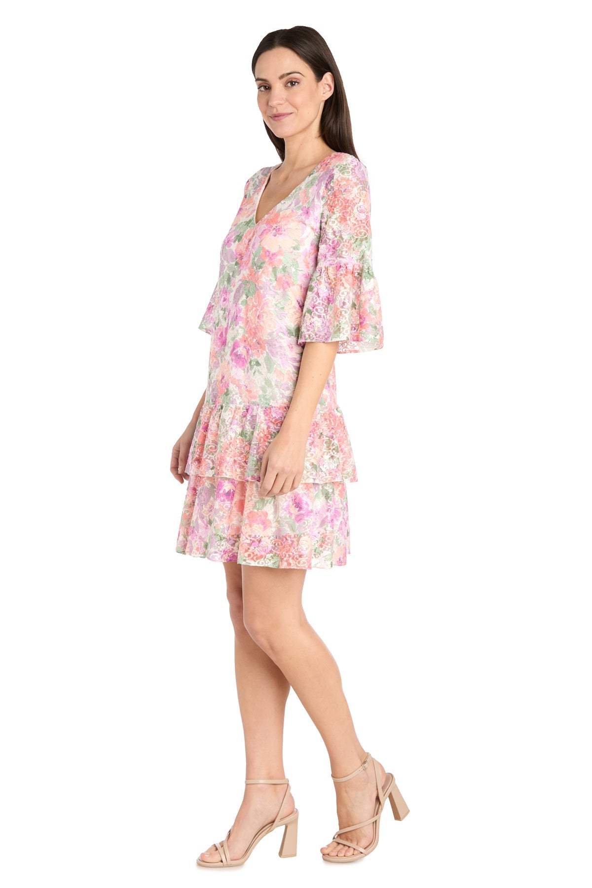 A woman poses in profile against a white background, wearing the R&M Richards 4107 Short Ivory Floral Party Dress with ruffled sleeves and a tiered skirt, styled casually with nude heeled sandals.